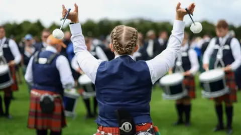 Getty Images Back of a female tenor drummers head. She is wearing a navy waistcoat and a red kilt. She is twirling tenor sticks in the air.