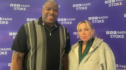 A woman with a grey hooded top over a green shirt stands next to a taller man with a black and grey shirt in front of a purple background with BBC Radio Stoke branding.