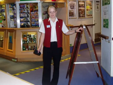 Sarah Clark A blonde haired woman in black trousers, white shirt and red waistcoast stands in front of a gift shop on board a ship