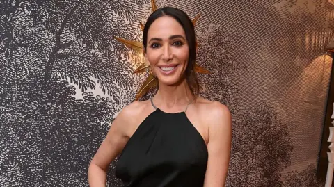 Dave Benett / Contributor/ Getty Images Lauren Cowell is wearing a black dress and is smiling at the camera. She has shoulder length brown hair and brown eyes. She is stood in front of a wallpapered wall that has a black and white drawing of a countryside scene on it.