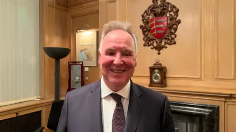 Simon Dedman/BBC Chris Whitbread wearing a suit and tie and standing in a wooden-panelled room with the Essex shield on a wall behind him, featuring the three Saxon seaxes.