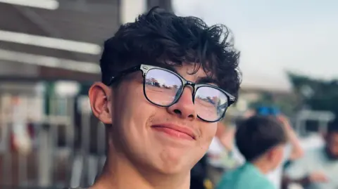 Michelle Lee Taylor Lee, a teenage boy with dark hair and glasses with dark frames, growing the beginnings of a moustache, smiling at the camera while sitting outside at a restaurant or cafe on holiday.