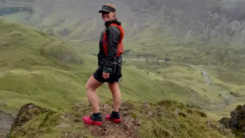 Becky Wightman Becky Wightman stands on top of the fell in black running clothes and wears an orange trail running backpack and pink shoes. She is in the Lake District. 