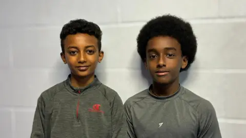 Schoolboys Yonatan Geremeskel and Mikey Abrehaley, both who have black hair and grey sportswear tops, smile at the camera against a white breezeblock wall. 