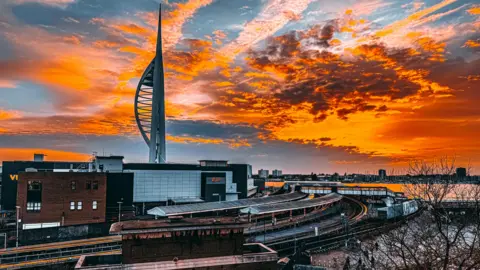 Lewis Jefferies The sky glows a dramatic bright orange colour over Portsmouth. There are dark clouds on the horizon. Below you can see the Spinnaker Tower and train station. You can see the glass building of a cinema and beyond the water of Portsmouth Harbour.