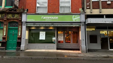 A deli stands on a gloomy street following a spell of rain. It has a green sign above the door and windows that reads "Farmhouse" and has a curtain down at the window, although the lights are on, as can be seen through the door.