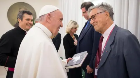 Osservatore Romano Pope Francis, dressed all in white, is holding the book and smiling as he talks to David Willey, who is also smiling.
