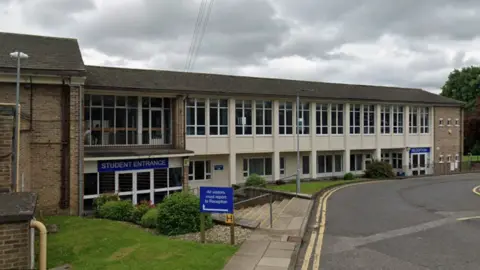 Google A two‑storey brick and concrete school or college building with large rectangular windows, separate doors marked “Student Entrance” and “Reception,” and a blue sign instructing visitors to report to reception, set beside a paved road with yellow lines on an overcast day.