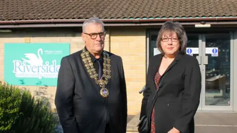 Image of Phil Exton and Kim Power looking into the photo. They are both wearing black jackets. Mr Exton is wearing a mayor's 'livery collar'. They are standing in from of Riverside Community Centre