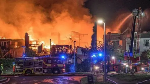 Fire engines with blue lights parked in front of a building consumed by fire. Smoke is pouring out the top and the area is glowing orange. Jets of water can be seen being sprayed at the fire.