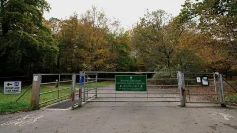 Peter Trimming/Geograph A gate with a sign on it which reads "Burnham Beeches National Nature Reserve". There is a smaller gate on the right for pedestrians and another small gate on the left with a cattle grid for cyclists. There is a sign on the left which reads "overflow car park". There is woodland in the background and it appears to be Autumn as there are lots of leaves on the ground.