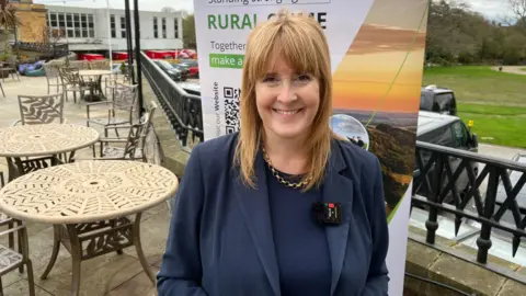 BBC/RICHARD EDWARDS Crime and policing minister Sarah Jones - pictured at today's national rural crime conference in York - is smiling at the camera. She is standing outside, with tables and chairs behind her, and is wearing a blue suit and a gold necklace. A Rural Crime Network banner is behind her.