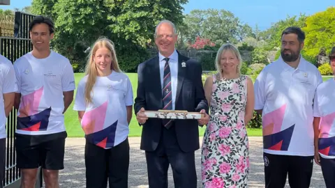 Group of people stand outside a gated entrance in a garden setting; several wear white “King’s Baton Relay Glasgow 2026” shirts while a central figure in a suit holds a decorated baton, with greenery, trees, and a large open gate behind them.