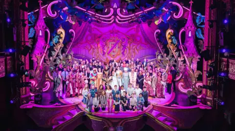 Danny Kaan The full cast of Sleeping Beauty on the stage at Wolverhampton Grand Theatre, which is all lit up in pink.