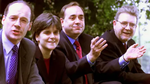 PA Media John Swinney and Nicola Sturgeon stand in a row with Alex Salmond and Michael Russell. They are all in business wear and are smiling and waving.