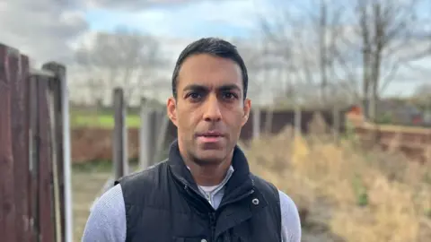 Simel Kara, wearing a black gilet jacket and a jumper, looks towards the camera. In the background the roughoutline of a fence can be seen at the back of the family property.