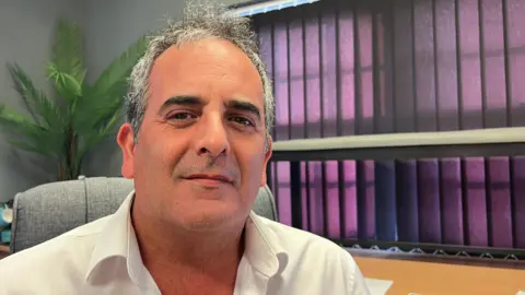 A man with short salt-and-pepper hair smiles as he sits on a grey chair at a wooden desk in an office with a potted green fern and purple blinds behind him. He is wearing a crisp white shirt. 