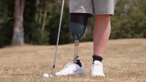 A close up of a man with a prosthetic leg stood next to a golf club and a golf ball