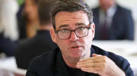 A close-up view of Burnham seated indoors, captured from the chest up. Burnham has short, neatly styled hair and is facing slightly toward the camera.
He is wearing dark rectangular glasses, a dark blazer, and a dark shirt. One hand is raised in front of the body, with the fingers held horizontally as if emphasizing a point while speaking. A ring is visible on one finger.

The photograph is sharply focused on the person in the foreground, while the background is heavily blurred. Several other people appear to be present in the background, but they are out of focus and their features are not clearly visible.