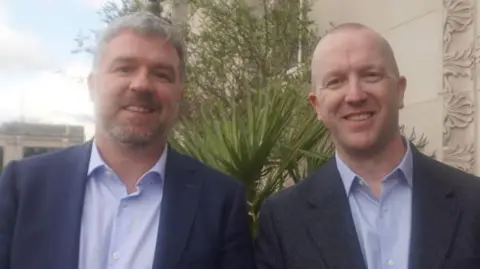 A man with grey hair and a beard is wearing a navy suit and pale blue shirt. He is standing next to a man with a bald heard in a dark suit with a pale blue shirt. Both are stood smiling in the street