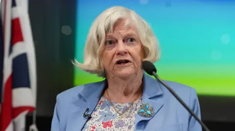 Anne Widdecombe speaking to a microphone wearing a light blue jacket and floral blouse. 