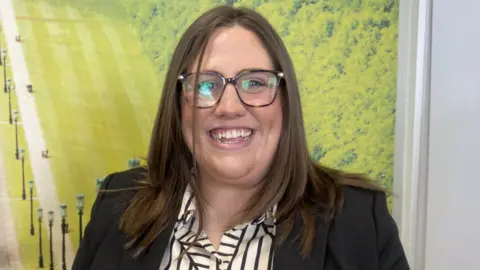 Julie Middleton has long brown hair and is wearing black-rimmed glasses.
She is wearing a striped white and black shirt with a black blazer and standing in front of a poster of the front gardens of Stormont