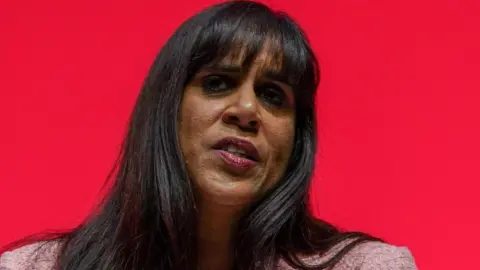 A woman with long dark hair is wearing a pink jacket. She is in front of a red background.