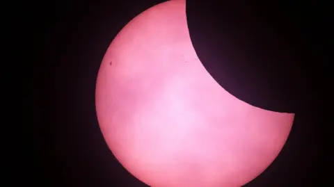 The top right corner of the sun has been obscured by the moon which appears black. The sun appears pink in the picture from St Martins in 2025.