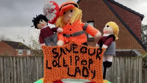 A knitted post box topper of a lollipop lady with two children and an orange sign saying 'save our lollipop lady'