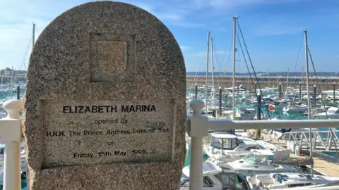 A granite marker with the following words imprinted: ELIZABETH MARINA, opened by HRH The Prince Andrew, Duke of York on Friday 15th May 1998. Behind the stone are many motor boats and yachts tied up to pontoons.  