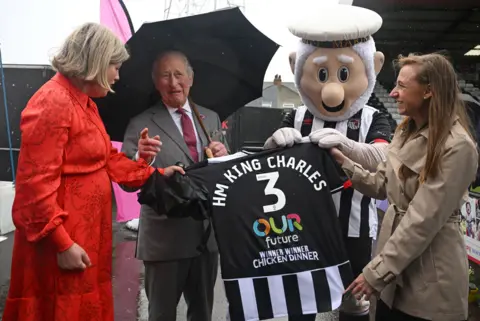 PA Media King Charles is presented with a Grimsby Town shirt with the lettering "HM King Charles 3 Our Future Winner Winner Chicken Dinner". The shirt is being presented by the club mascot The Mighty Mariner. There are two women also involved in the presentation. 