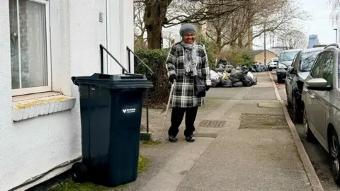 Gabriel Bononi A woman wrapped up in a big coat and cosy hat stands on a pavement. IN front of her is a black wheelie bin in front of a white house. Behind her is a pile of about 20 rubbish bags