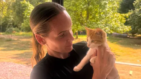 Natalie Bhangal A woman holding a ginger and white kitten closely in front of a window onto a garden on a sunny day. She is wearing a black top and has long brown hair tied back in a ponytail. 