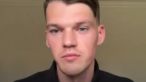 A headshot of Ben Francis with short dark hair. He is looking at the camera, whilst wearing dark coloured clothing, in front of a neutral background.
