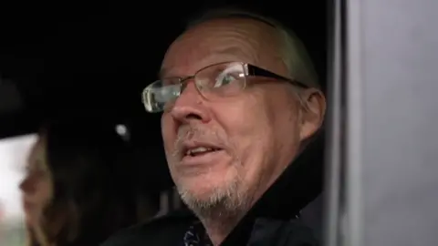 A middle-aged man with grey hair, short grey beard and glasses. He is sitting in the passenger seat of a car looking to the left. In the background is a blurred figure