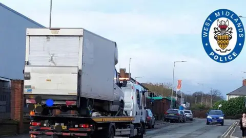 A street with industrial units on the left side and a residential house surround by a hedge on the left. In the foreground there is a white truck with a trailer that has a white van on the back. In the background there are four cars parked on the left and one blue car driving towards the camera on the right. A woman is just visible on the right of the frame