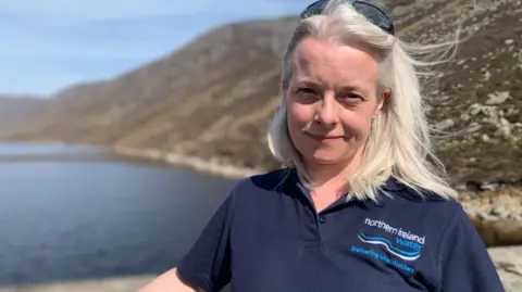 A woman with shoulder length fair hair is standing in front of a wall with water behind her. Sunglasses are pushed up on the top of her head and she is wearing a blue t-shirt with the Northern Ireland Water insignia on it.