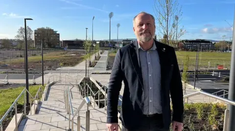 A man stands in the middle of the frame, wearing a blue shirt and black jacket. He is smiling at the camera. Behind him are stairs leading to the walkway and green fields.