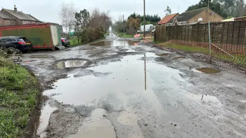 John Devine/BBC A view along a pothole-filled road with lots of standing water, no lines on the road or street lights. Some buildings and vehicles can be seen parked off the road on either side. There is a reflection of a telegraph pole in the main puddle in the foreground. The appears to be some sort of shipping container or lock-up on the left-hand side of the road.