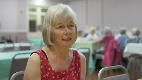 A woman with blonde hair in a bob sits in a community cafe with others in the background. She wears a red dress with floral print and has a serious expression on her face.