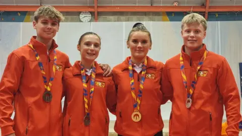 Helen Duggan Four young Manx athletes, two women and two men, wear red jackets and stand with arms around each other. They each have different coloured medals around their necks.