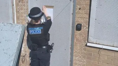A female police office sticking a bit of paper to a boarded up door. The door is covered in a metal grate material, as are the windows on either side of the light brick building. The police officer has brown hair which is in a bun and a black police hat on. She is wearing all black and a vest which has police written in white in a blue box on the back. Her belt has handcuffs attached to it.