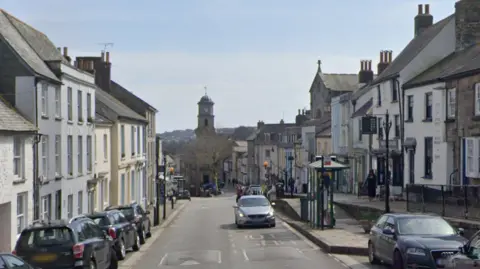 Lower Market Street in Penryn on a bright day. The road has road humps near a bus stop and there are cars parked on both sides. At the end of the street is a clock tower with a rounded dome and cross. The buildings lining the pavements on both sides are three and four storeys and some have shop windows on the ground floor.