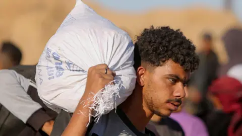 A Palestinian man carries a white aid sack from the GHF over his right shoulder  in Khan Younis on May 29, 2025. He has curly hair, a moustache and a very light beard.