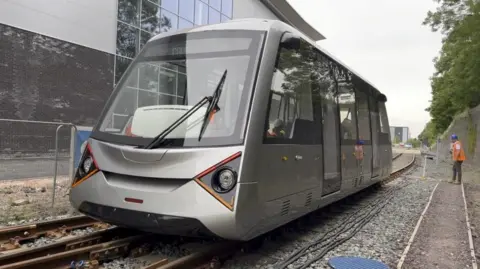 A modern silver train carriage on a length of track. A man in orange hi-vis jacket is standing next to it.