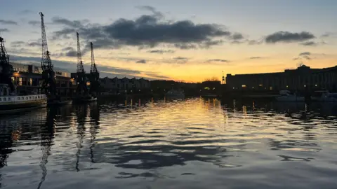 A view from the Prince Street Bridge looking towards the MShed Museum on Bristol Harbourside. It is taken at dusk and the water is dark and the sky is tinted orange. The cranes of the MShed are silhouetted dark against the sky as are the buildings of the Lloyds Ampitheatre. There are small ripples on the water but no boats