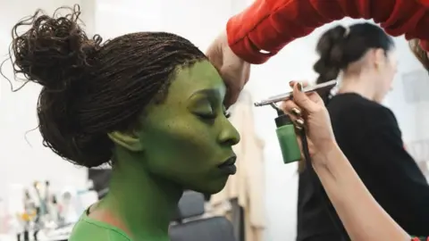 Cynthia Erivo sat in a make-up chair in a make-up trailer on the set of Wicked. She has her eyes closed and is being painted green by someone who is using an air brush machine. 