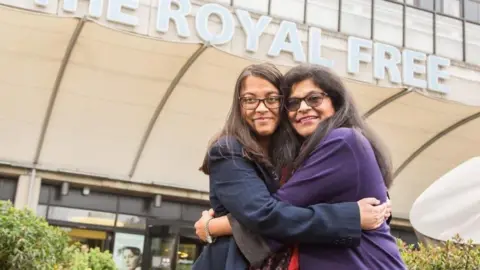 Prafula Shah Two women hug each other outside the Royal Free Hospital. Both women have long dark brown hair and brown eyes and are wearing glasses. The younger woman on the left is wearing a dark blue suit jacket. The older woman on the right has a purple jacket on.