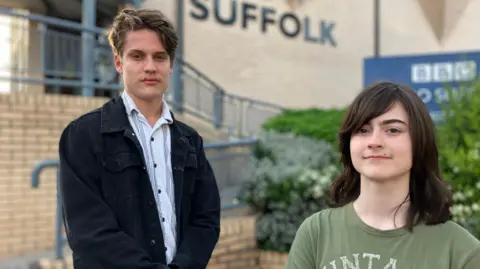 Hux Josecelyne and Jett Aiello stand in front of the BBC Radio Suffolk building. Hux, on the left, has a corduroy black shirt on unbuttoned, and Jett has long dark hair and is wearing a green t-shirt. 
