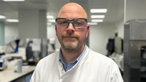 Jesse Peterson is standing in a lab with blurred machinery in the background. He is wearing a white lab coat over a blue shirt, along with safety goggles. He is bald with a short cropped beard and is half smiling at the camera.